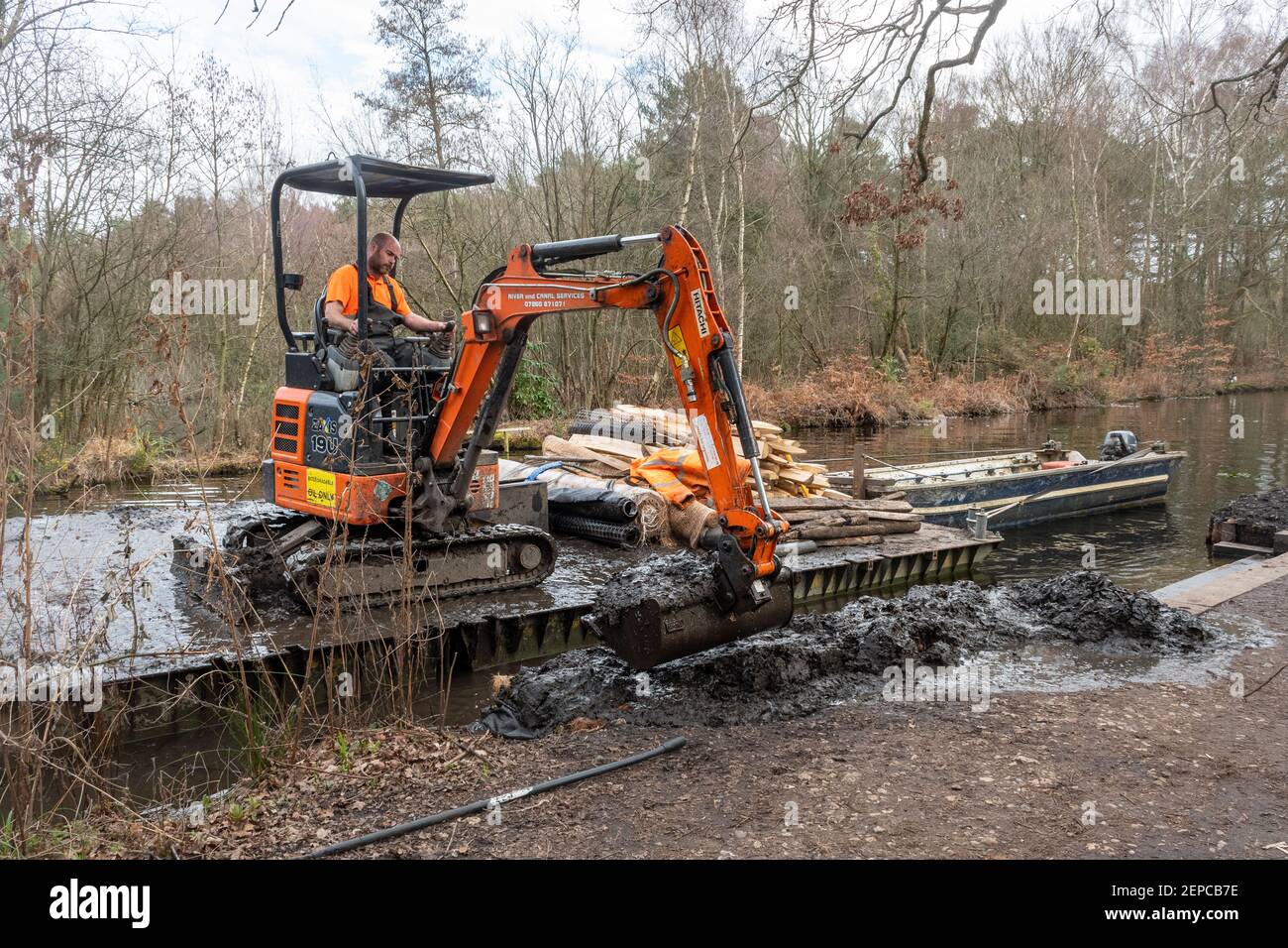 Hombre trabajando en una excavadora o excavadora en un pontón de trabajo flotante reparando un banco de canales erosionado en el Canal Basingstoke en Surrey, Reino Unido Foto de stock