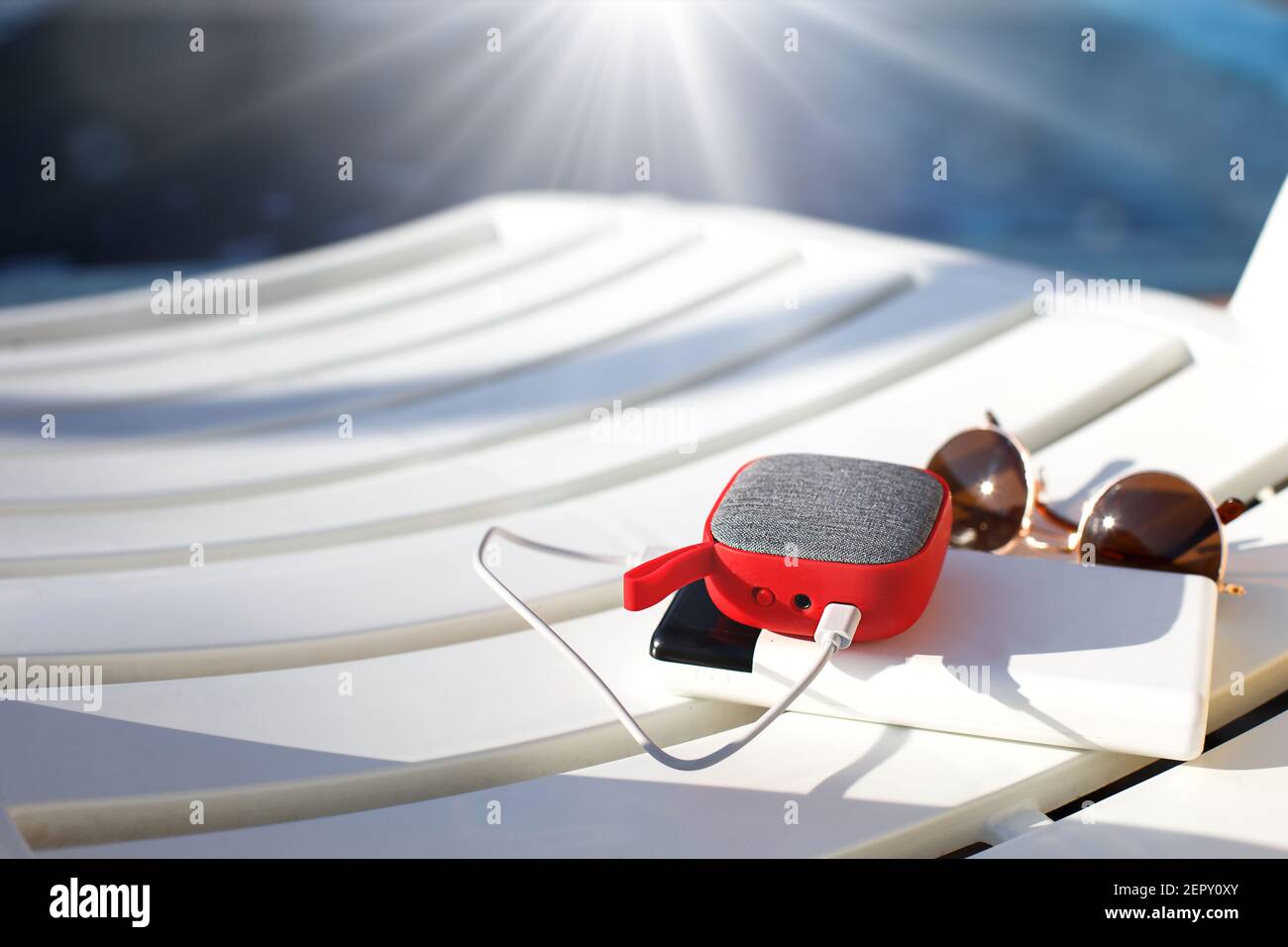 El altavoz portátil de música roja se carga desde la fuente de alimentación  a través de usb en una silla de cubierta cerca de la piscina. El concepto  está siempre en contacto,