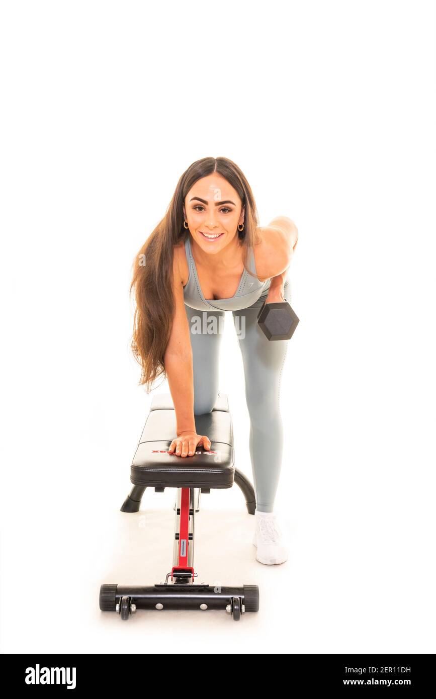 Retrato vertical de una joven usando una campana tonta y un banco de gimnasio para hacer ejercicio, aislado sobre un fondo blanco. Foto de stock