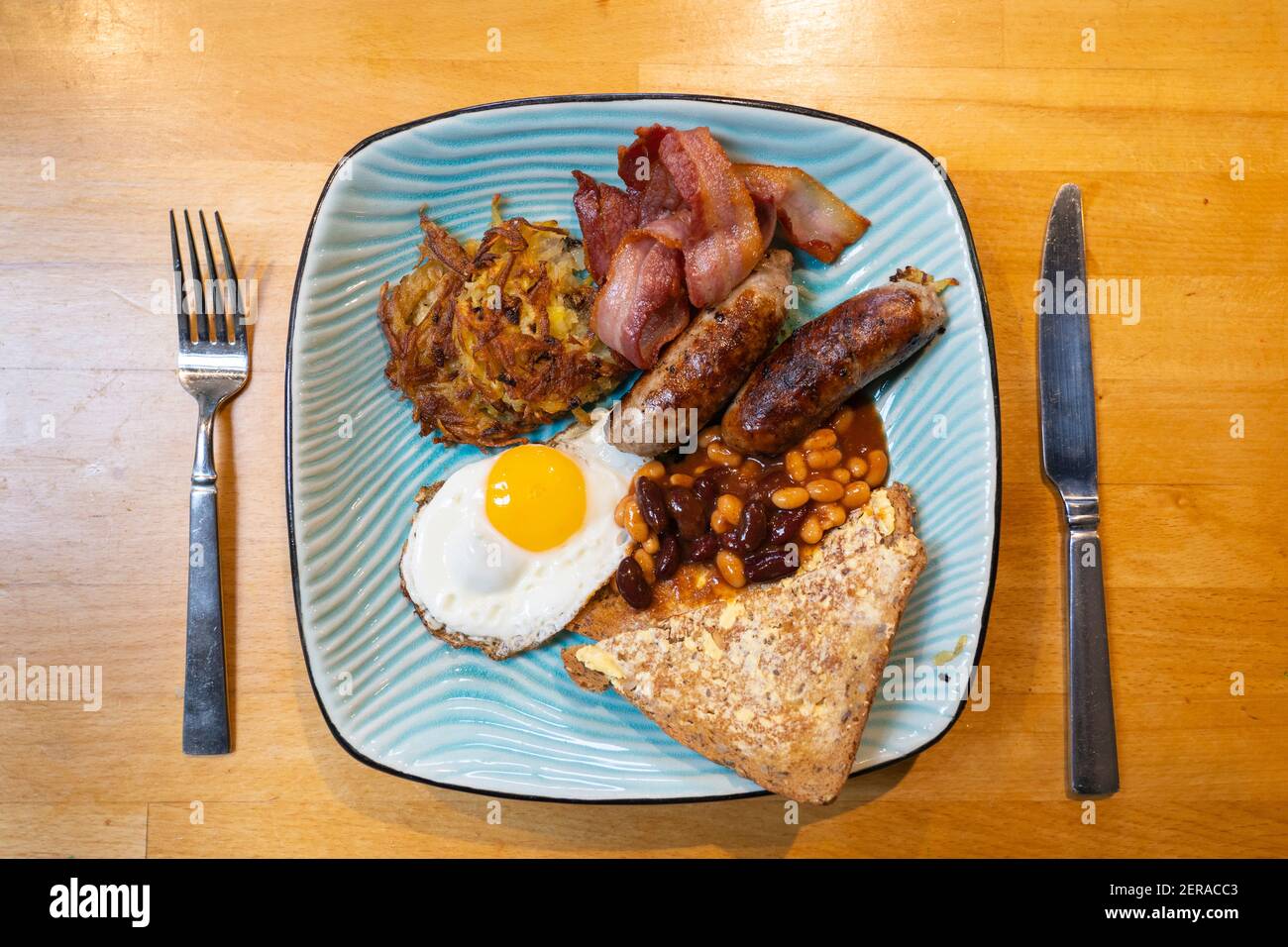 Un desayuno inglés tradicional servido en un plato azul con salchichas, un huevo frito, rashers de bacon, un hash brown, frijoles y rebanadas de pan tostado Foto de stock
