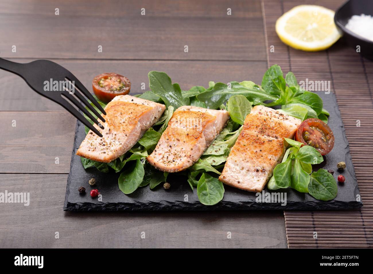 Tenedor pricando filetes de salmón sobre plato de pizarra sobre fondo de mesa de madera. Foto de stock