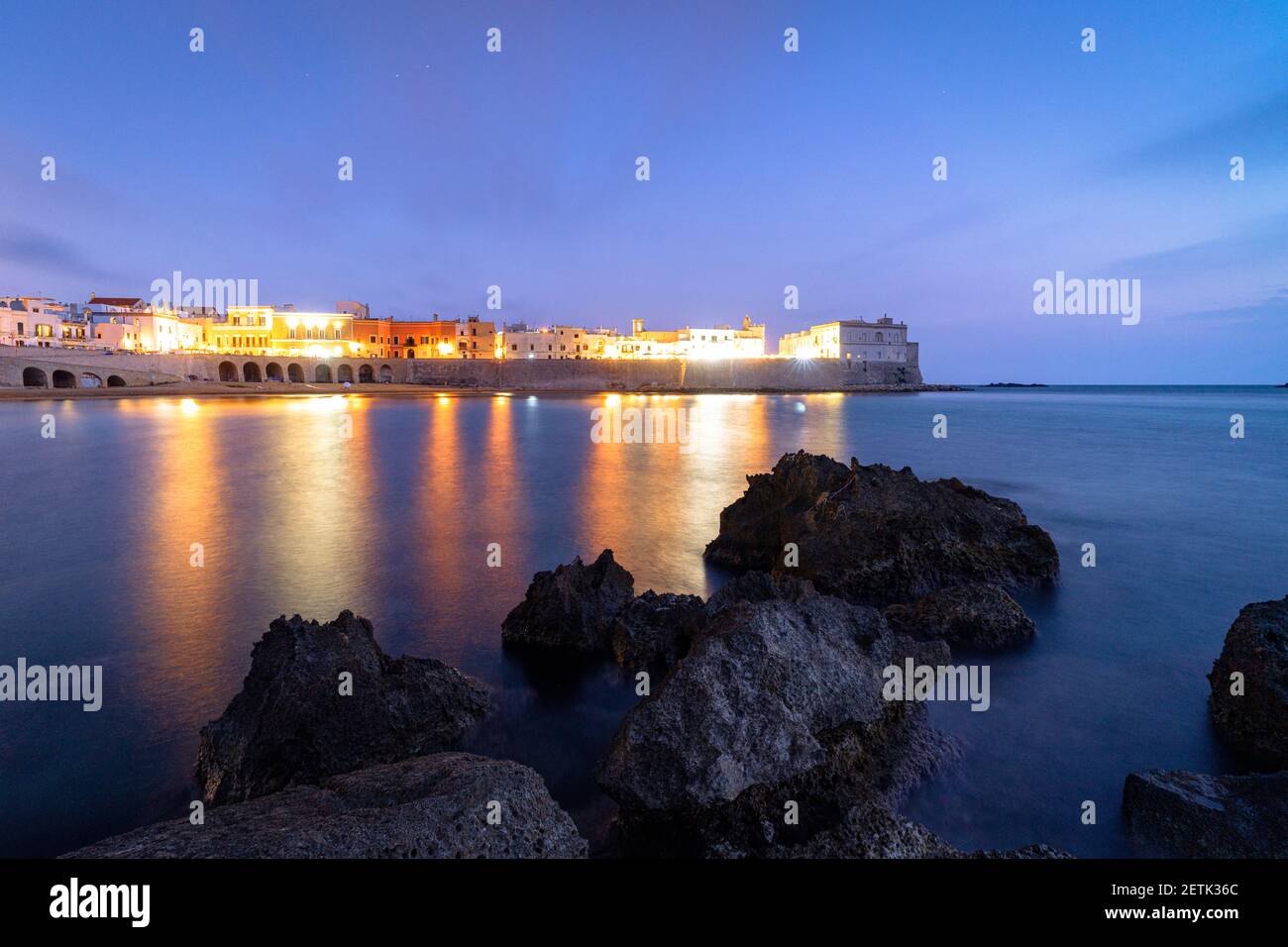 Edificios iluminados de Gallipoli reflejados en el mar azul al atardecer, provincia de Lecce, Salento, Apulia, Italia Foto de stock