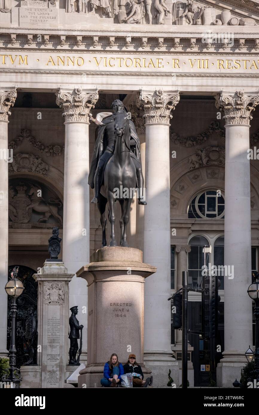 Dos jóvenes descansan bajo la estatua de la estatua ecuestre del duque de Wellington, que se encuentra frente a la Bolsa Real en la ciudad de Londres, el 1 de marzo de 2021, en Londres, Inglaterra. Diseñado por Sir William Tite en 1842-1844 e inaugurado en 1844 por la Reina Victoria, Royal Exchange es el tercer edificio del tipo erigido en el mismo sitio. El primer intercambio erigido en 1564-70 por el señor Thomas Gresham fue destruido en el gran incendio de 1666. Su sucesor, de Jarman, también fue quemado en 1838. El edificio actual es el grado 1 enumerado y cuesta alrededor de £150,000. Foto de stock