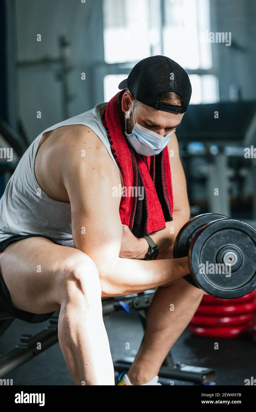 Hombre con máscara haciendo ejercicios de bíceps con pesas en el gimnasio.  Sentado en un banco. Covid19, coronavirus. Concepto de salud y bienestar  Fotografía de stock - Alamy