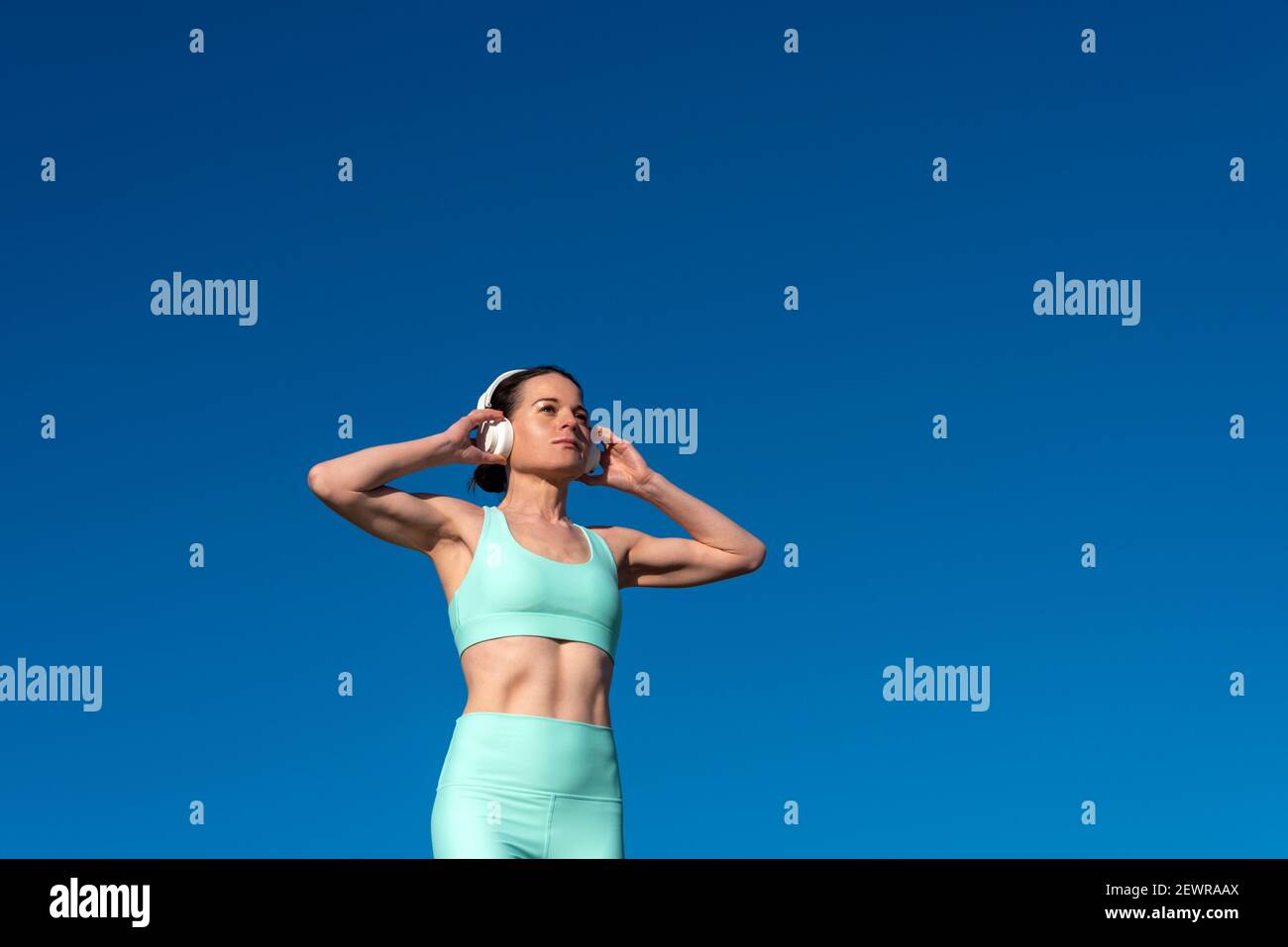 Mujer deportiva con sujetador deportivo y auriculares mientras hace ejercicio al aire libre con un cielo azul. Foto de stock