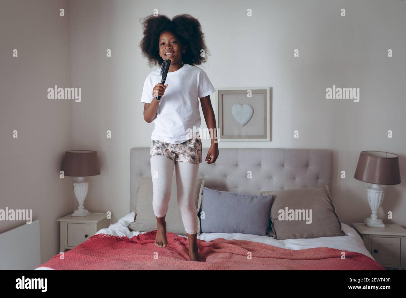 Niña afroamericana de pie en la cama sosteniendo el cepillo de pelo fingiendo canten Foto de stock