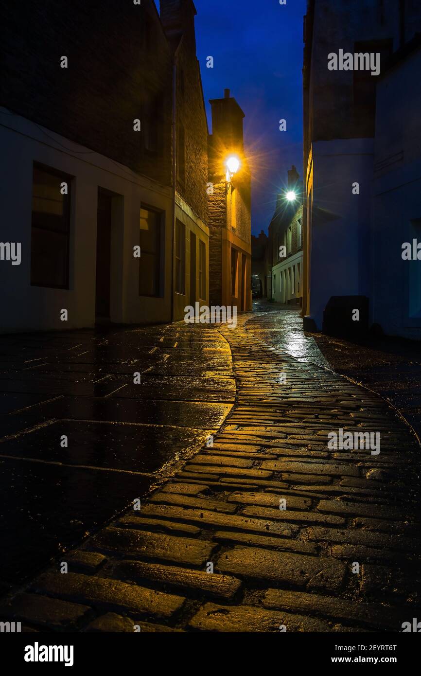 Curva calle estrecha entre las casas tradicionales escocesas en Orkney Ciudad Stromness por la noche con luces de calle Foto de stock