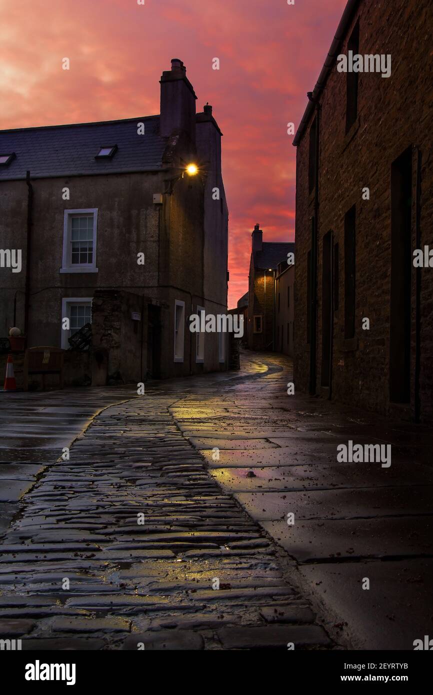 Amanecer cielo rosa con calle estrecha en la ciudad escocesa Orkney islas con curva estrecha calle de guijarros Foto de stock