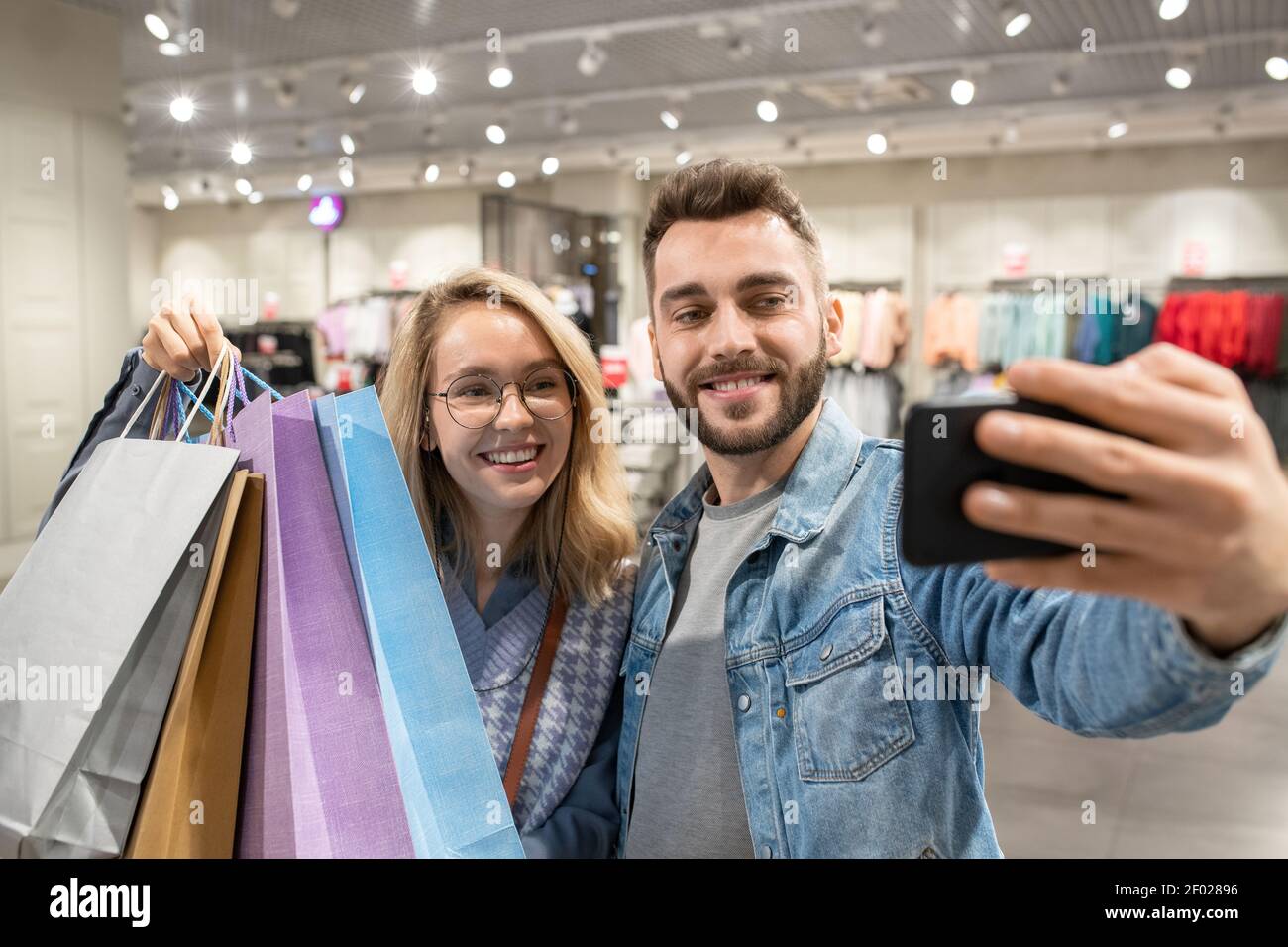 Joven feliz pareja con bolsas de compras haciendo autorretrato teléfono móvil en la tienda Foto de stock