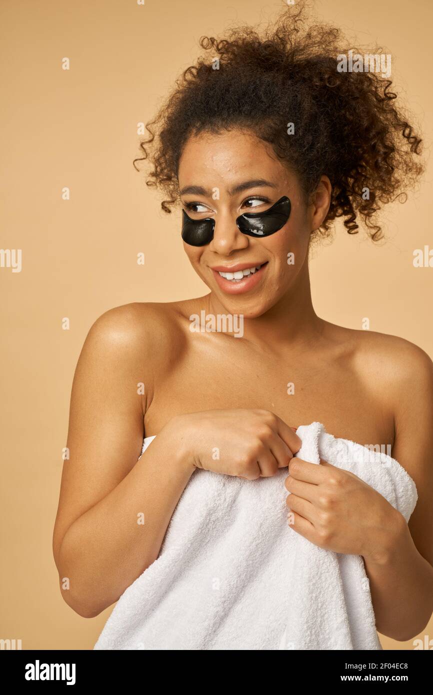Concepto de personas, belleza e higiene. alegre mujer afroamericana usa  gorro de baño, envuelto en una toalla, se aplica parches debajo de los ojos  después de tomar una ducha, se preocupa por