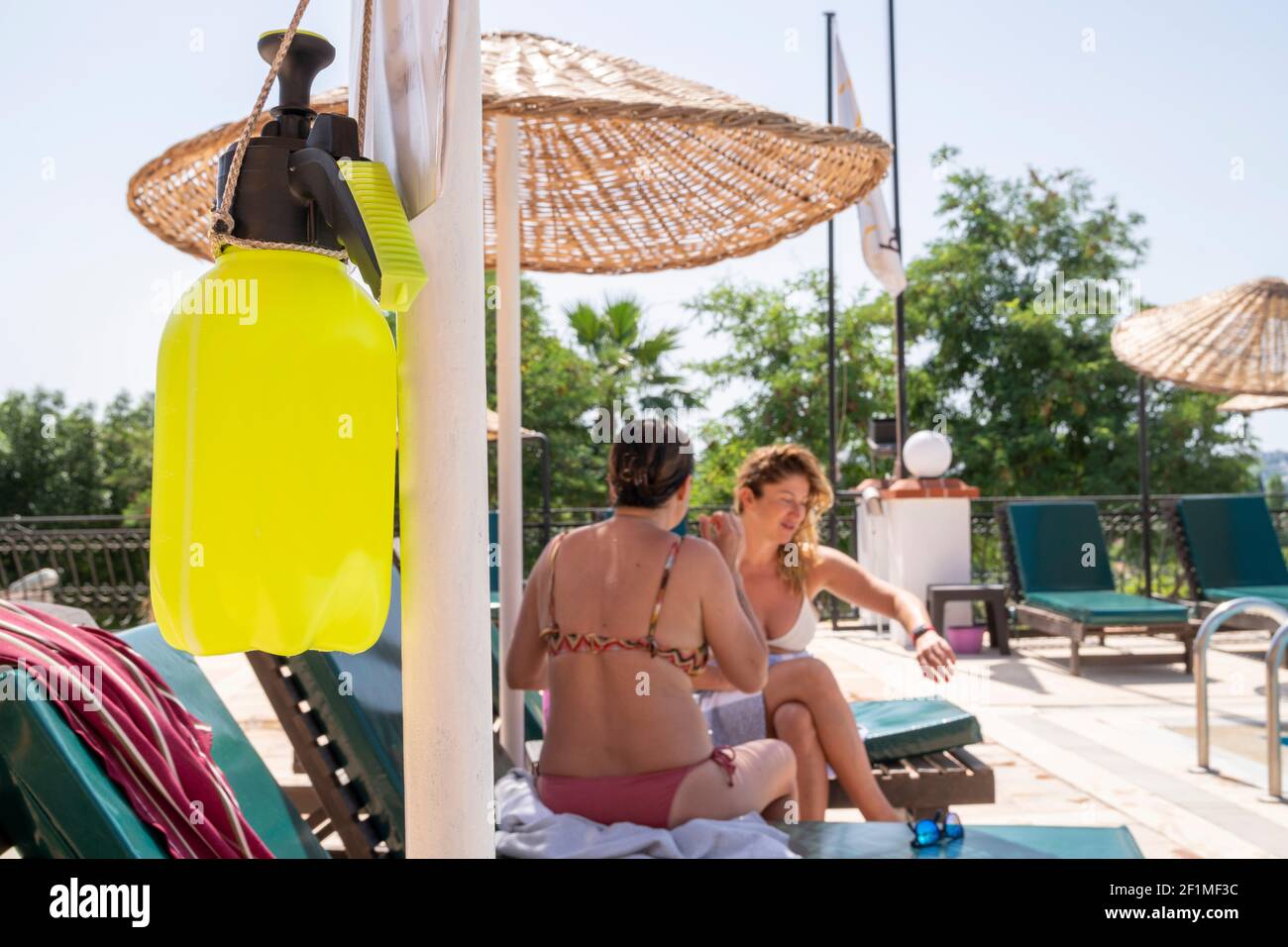 Verano 2021 - Nuevo normal. Desinfectante a base de alcohol contra Covid-19 colgado en un poste en la zona de la piscina. Dos mujeres sentadas en las tumbonas y hablando Foto de stock