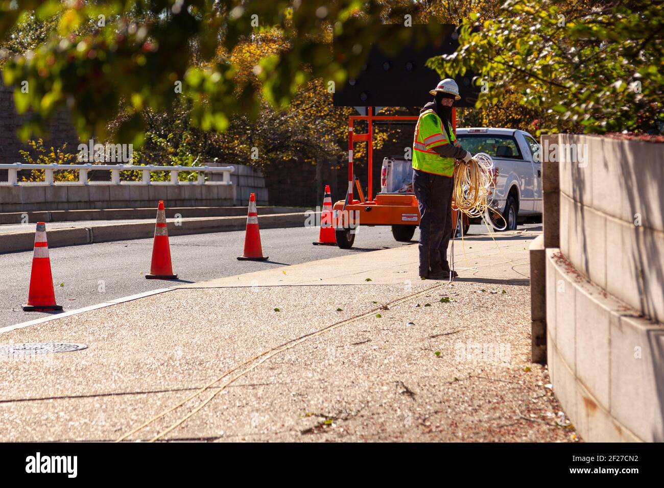 Washington DC, EE.UU. 11-02-2020: Un técnico de electricidad que lleva chaleco reflectante y casco está instalando el cable en un lado de la carretera. Hay conos a des Foto de stock
