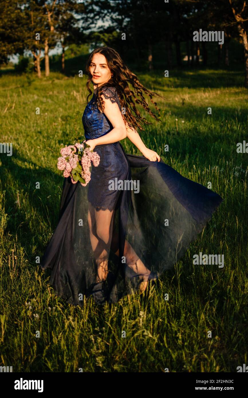 Fiesta de graduación prom de cuento de hadas concepto. Hermosa mujer joven  morena en vestido de gala azul con ramo de lila en el fondo de la  naturaleza. Romántico al aire libre