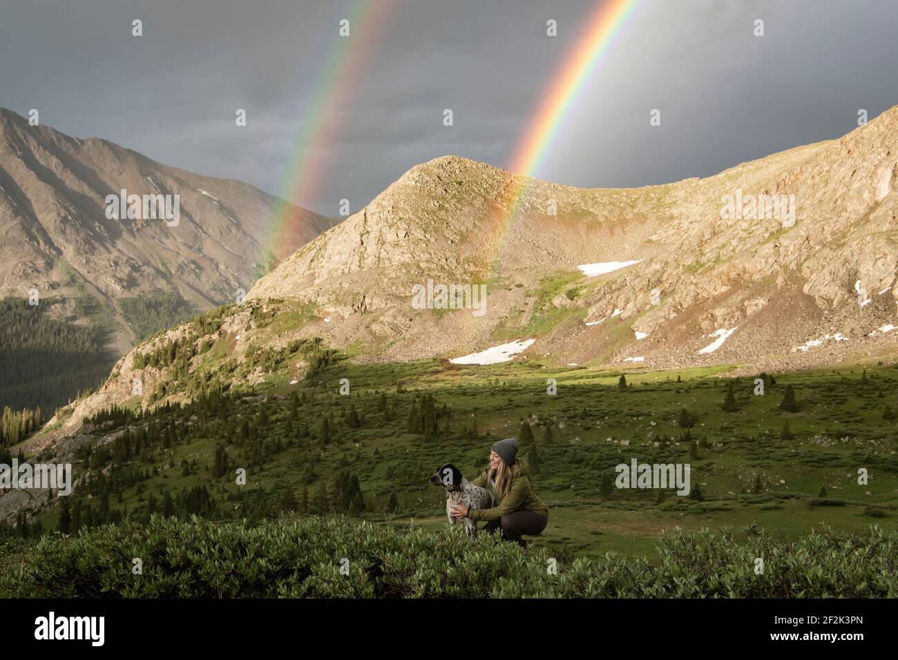 Mujer acariciando perro mientras doble arco iris y montañas en el fondo Foto de stock
