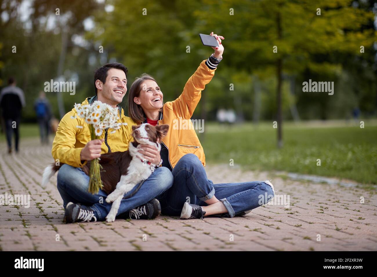 joven mujer caucásica tomando selfie con su novio y su perro en un regazo, sentado en un camino en el parque Foto de stock