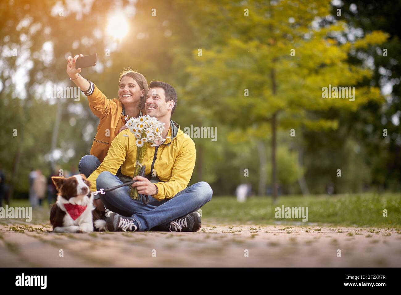 pareja caucásica con un perro sentado en un camino en el parque con las piernas cruzadas, tomando selfie con su perro Foto de stock