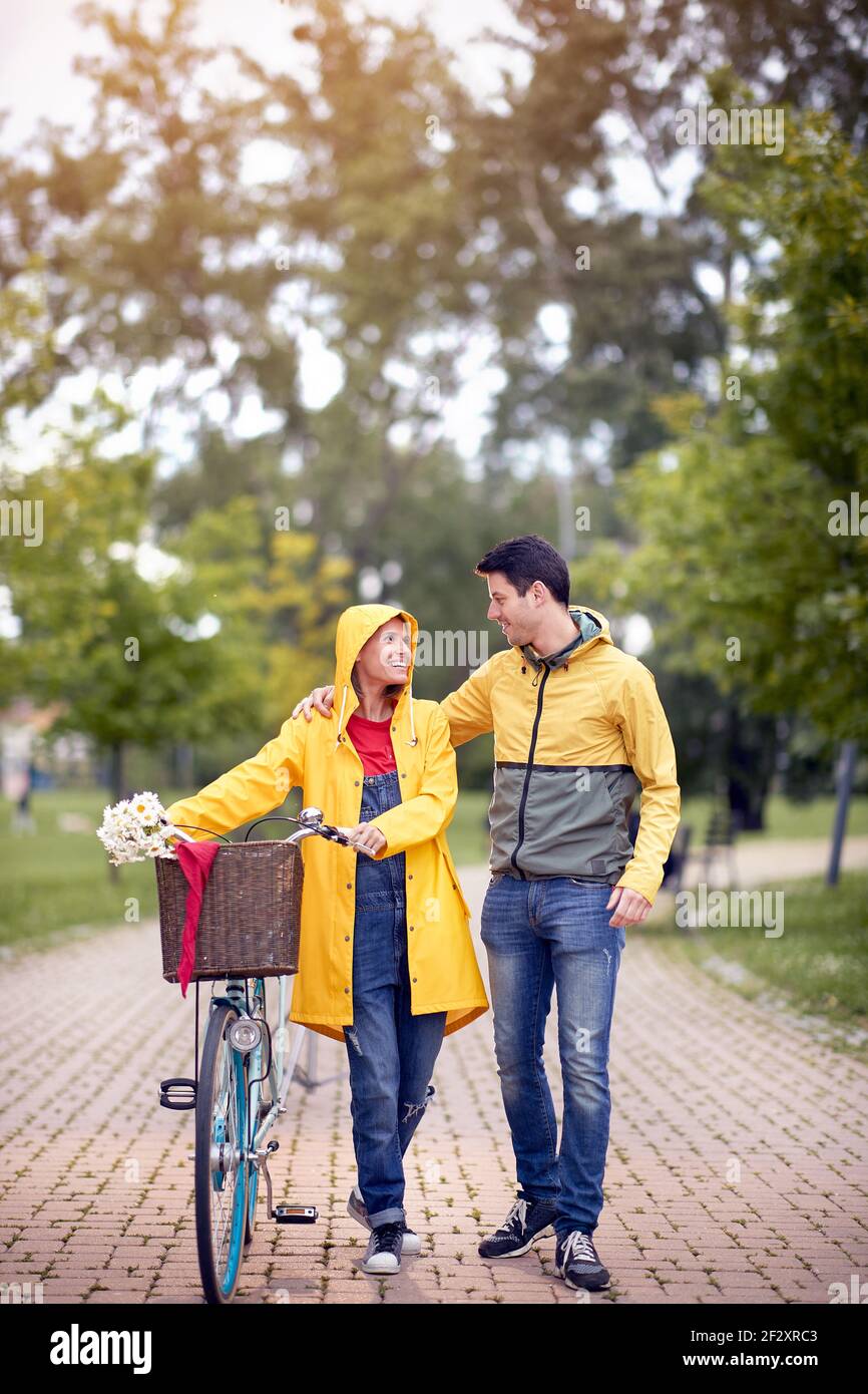 imagen vertical de hermosa mujer mirando con amor y afecto un joven caucásico abrazándola mientras empuja la bicicleta un día lluvioso en el parque Foto de stock