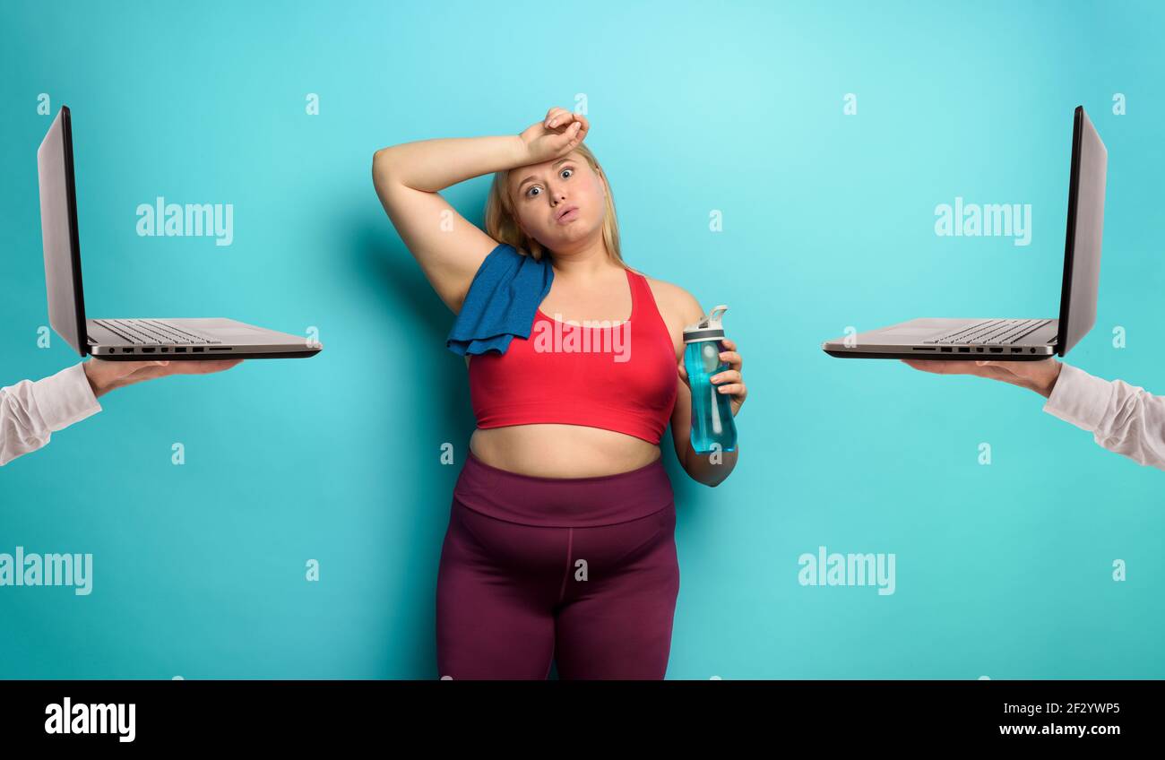 Chica gorda hace gimnasio en casa de forma remota con portátil. Expresión cansada. Fondo cian Foto de stock