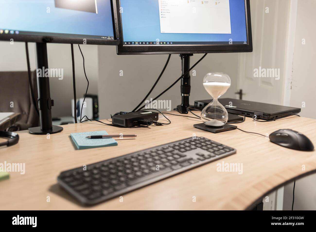 Una vista de cerca de un escritorio de una estación de trabajo con monitores, teclado, ratón, portátil, estación de acoplamiento y un temporizador de arena utilizado como técnica de enfoque. Foto de stock