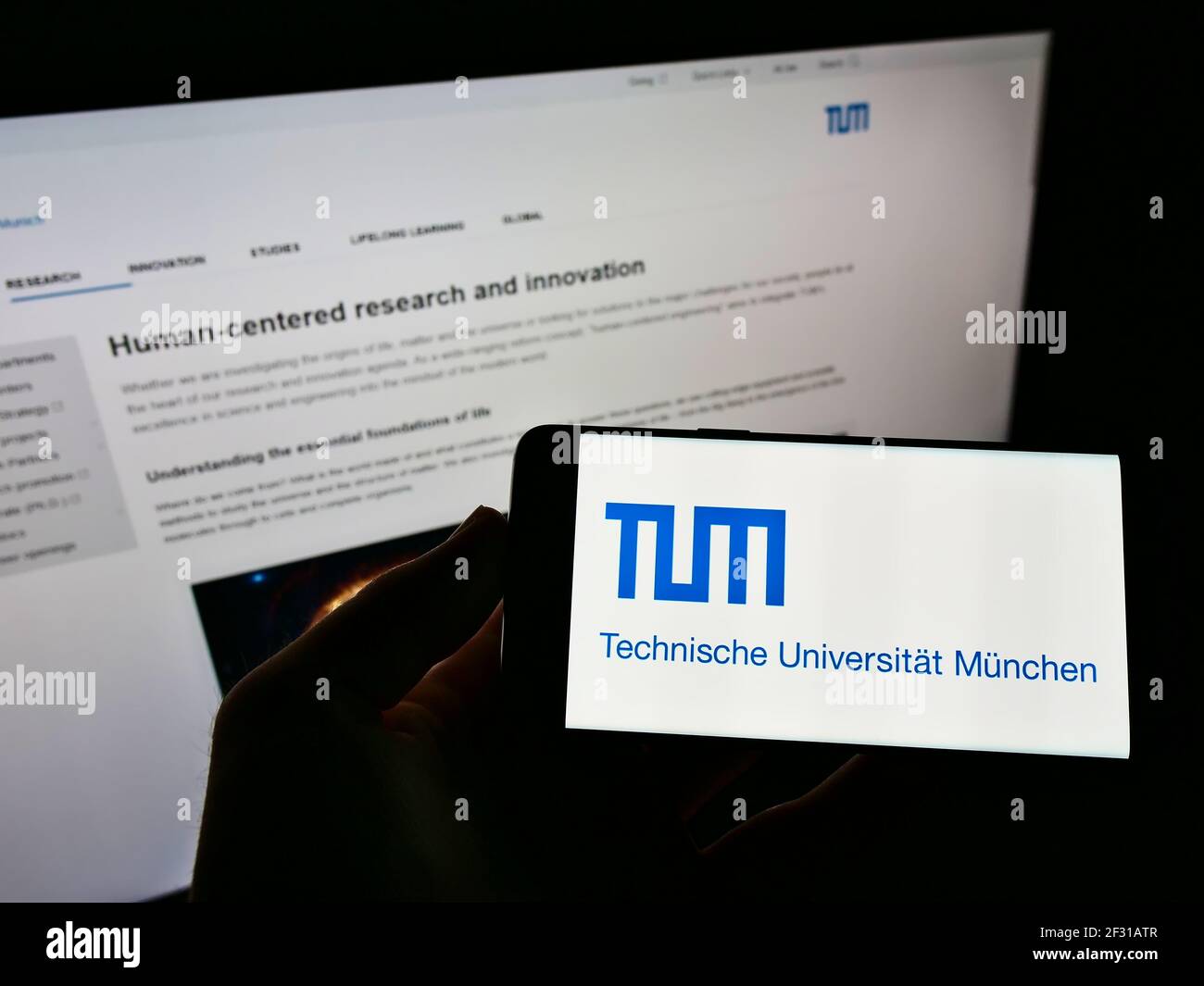 Persona que sostiene el smartphone con el logotipo de la universidad alemana Technische Universität München en la pantalla frente a la página web. Enfoque en la pantalla del teléfono. Foto de stock