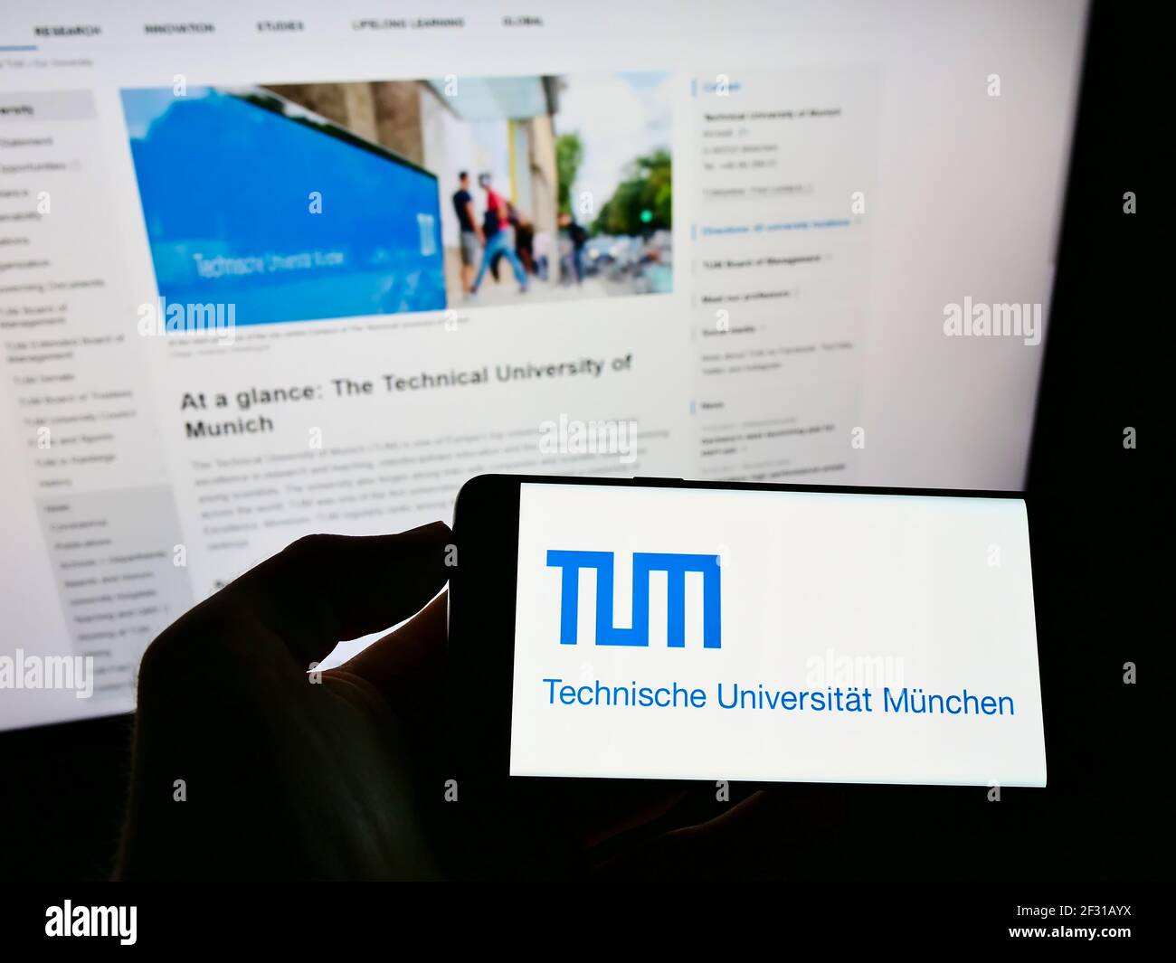 Persona que sostiene el smartphone con el logotipo de la institución educativa Universidad Técnica de Munich en la pantalla frente a la página web. Enfoque en la pantalla del teléfono. Foto de stock