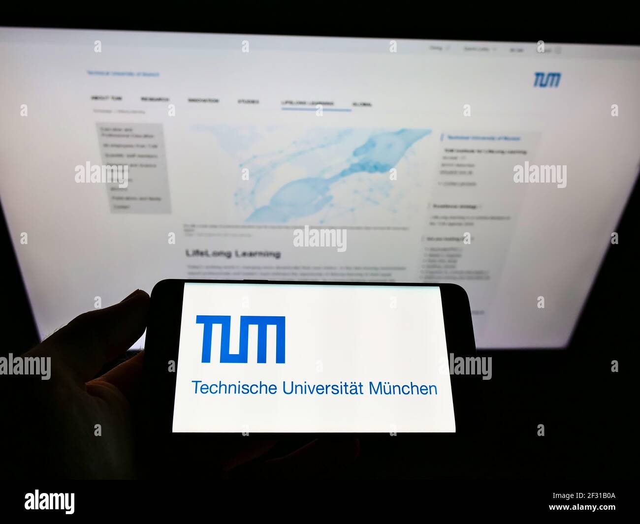 Persona que sostiene el teléfono celular con el logotipo de la institución educativa alemana tu Munich en la pantalla frente a la página web. Céntrese en la pantalla del teléfono móvil. Foto de stock