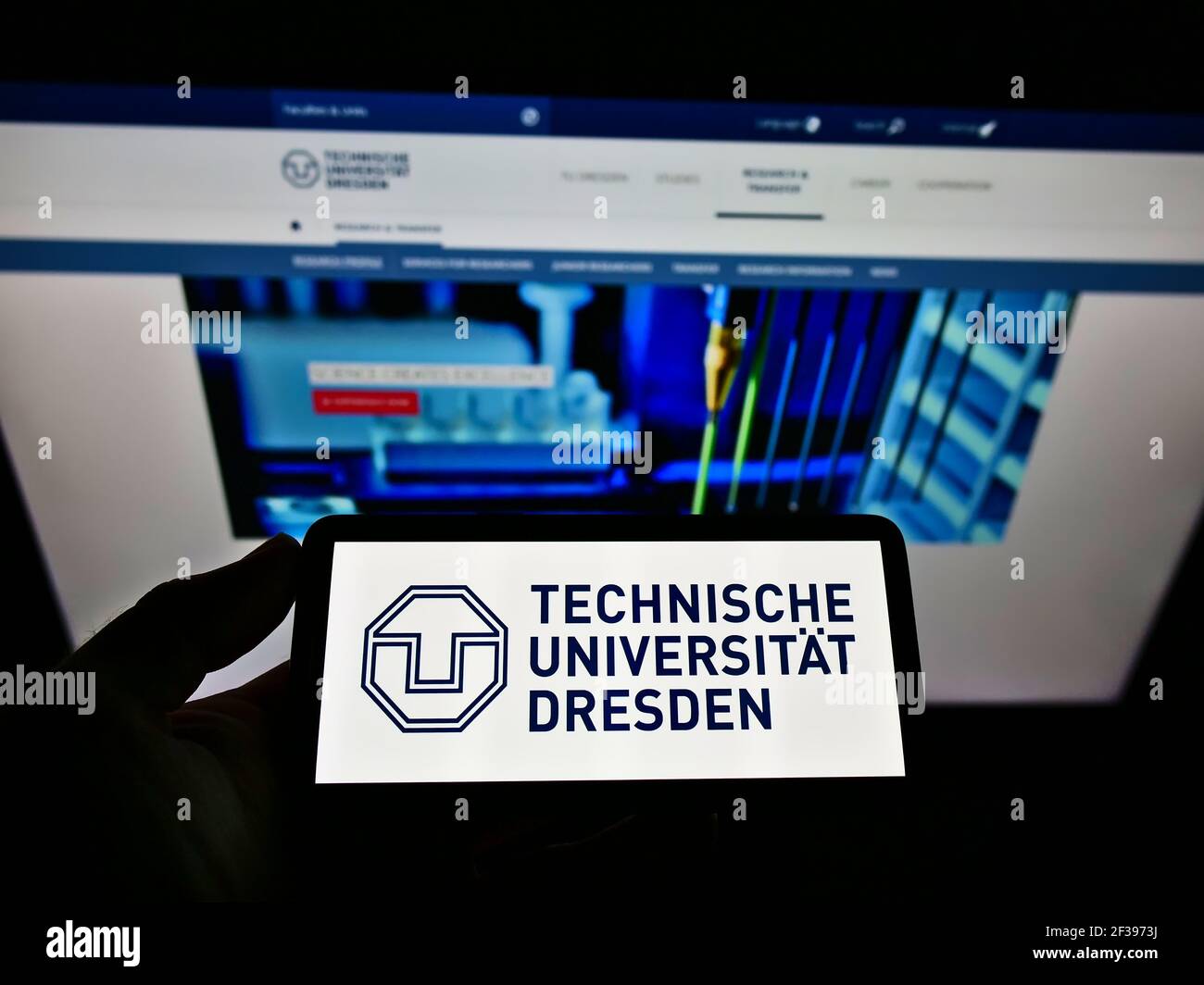 Persona que sostiene el teléfono móvil con el logotipo de la Universidad Alemana de Tecnología de Dresden en la pantalla frente a la página web. Enfoque en la pantalla del teléfono móvil. Foto de stock