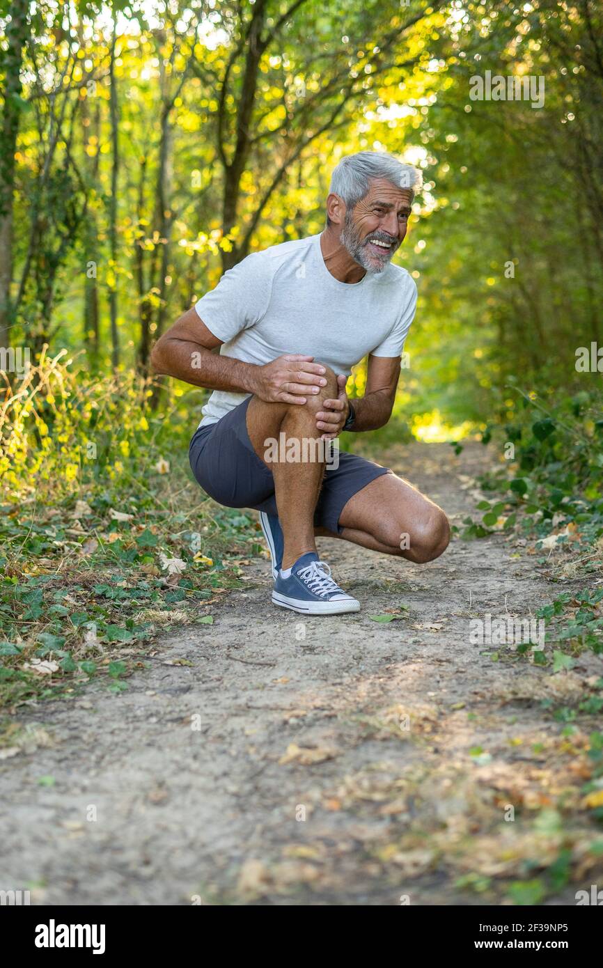 Hombre maduro que sufre de dolor de rodilla mientras se agachaba en el bosque Foto de stock