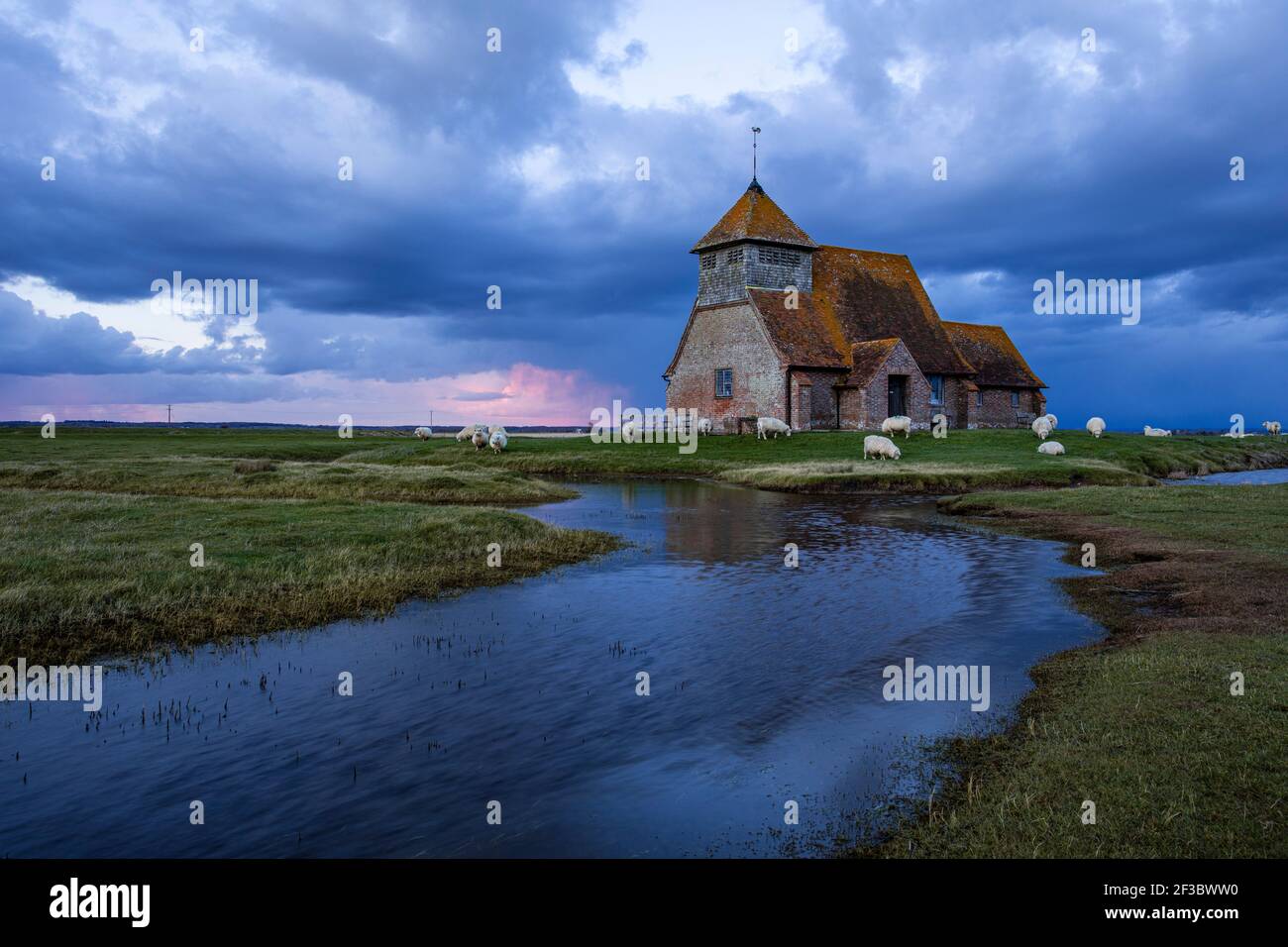 Iglesia de Santo Tomás Becket cerca de Fairfield en el pantano Romney en Kent sur este de Inglaterra, nubes de tormenta se reúnen durante la hora azul y pastoreo de ovejas Foto de stock