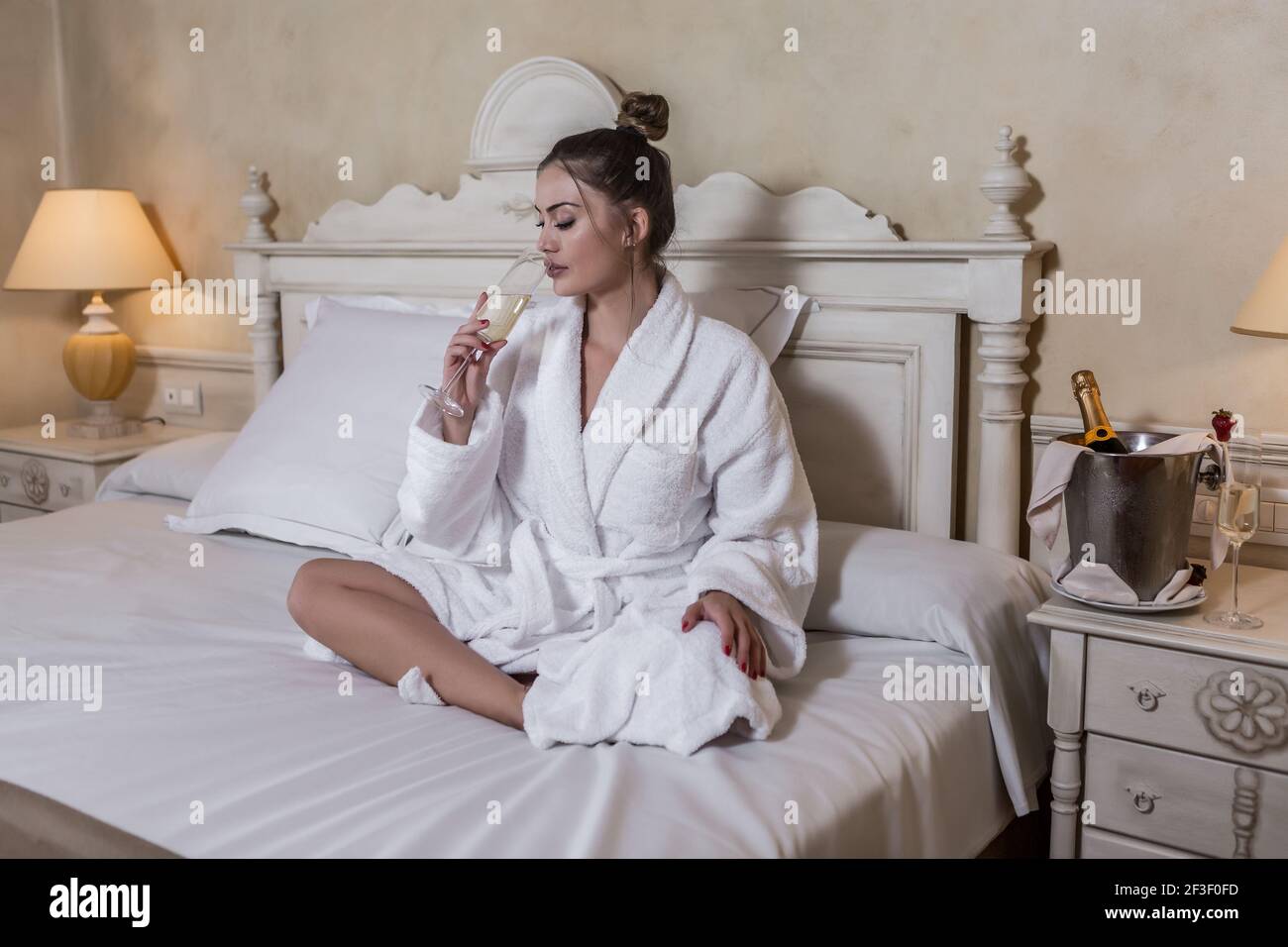 recoger escucha Arne Bonita señora en un albornoz blanco cerrando los ojos y bebiendo champán  mientras se sienta en una cama blanda en una habitación de hotel de lujo  Fotografía de stock - Alamy