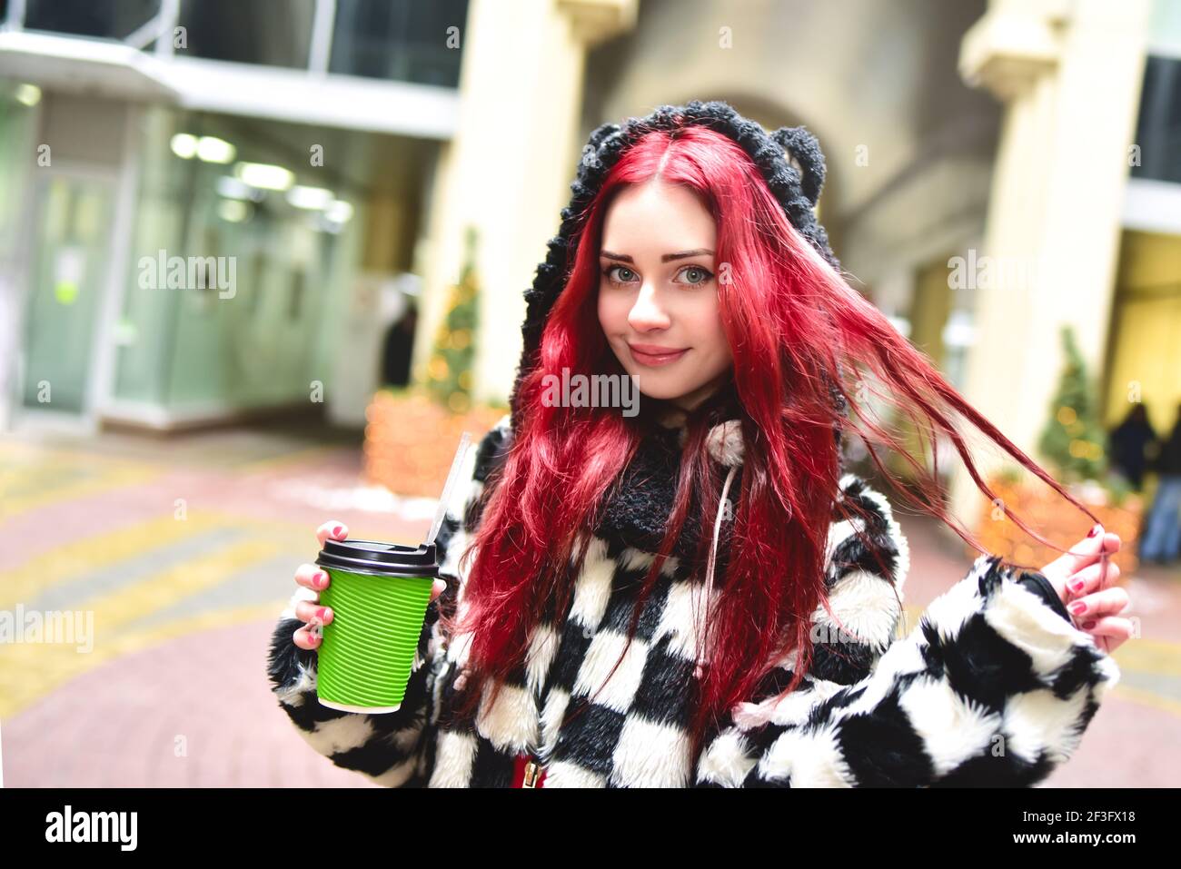 Cerca del retrato de una niña adolescente con pelo rojo en ropa caliente de pie afuera en un día frío con una taza de café en sus manos y mira en la cámara. Foto de stock