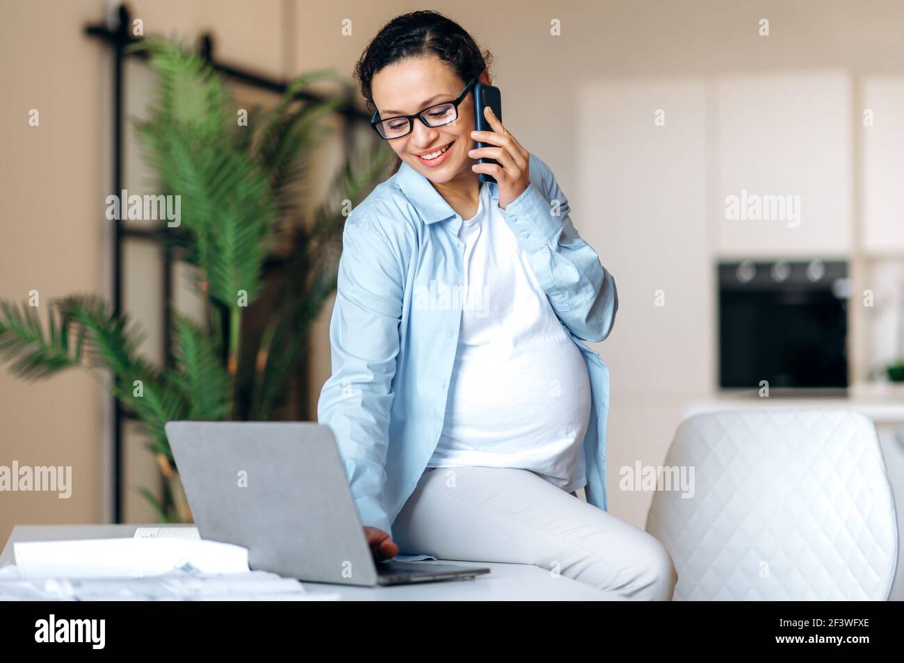 Alegre embarazada mixta raza adulta mujer en elegante ropa formal está  sentado en su escritorio de trabajo, teniendo agradables usos de  conversación celular y mirando el portátil, sonriendo Fotografía de stock -