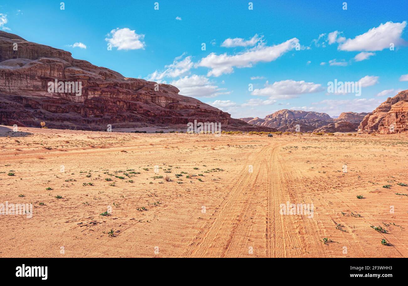 Macizos rocosos en el desierto de arena roja, cielo nublado brillante en el fondo, pequeño vehículo 4WD y camello a distancia - paisaje típico en Wadi Rum, Jordania Foto de stock