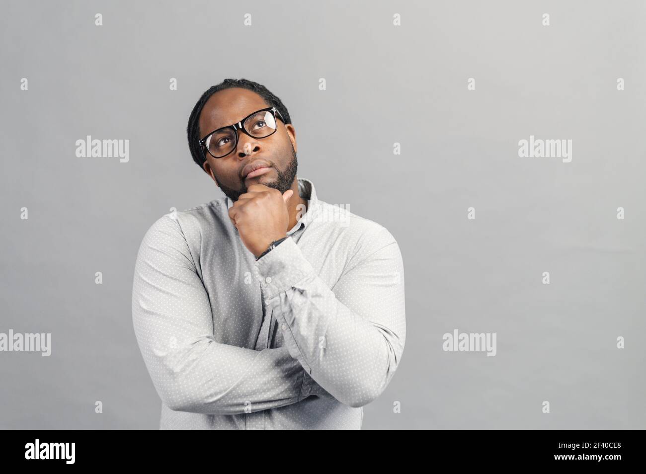 Hombre afro-americano pensativo con gafas con estilo y camisa elegante sport  buscando nuevas ideas e inspiración, sostiene la barbilla con una cabeza,  se ve, perdido en pensamientos, aislado en gris Fotografía de