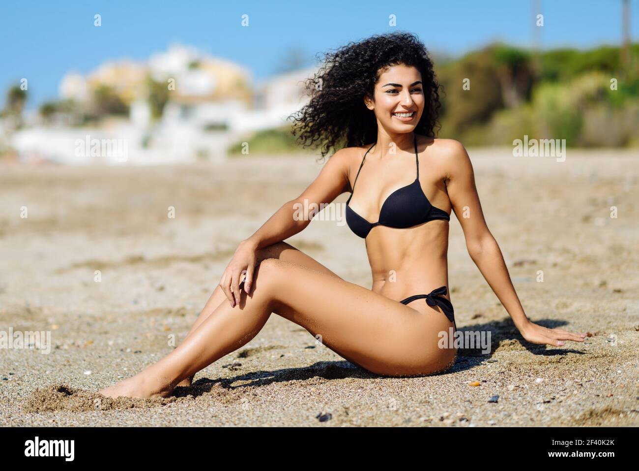 Joven mujer árabe con hermoso cuerpo en traje de baño sentado en la arena  de la playa. Mujer sonriente con peinado rizado largo usando bikini negro. Mujer  árabe con cuerpo hermoso en