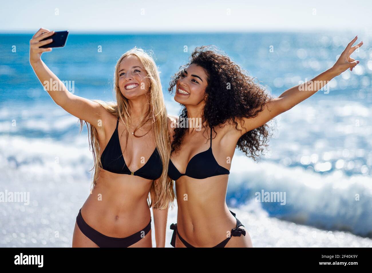 Dos mujeres jóvenes tomando fotografía selfie con teléfono inteligente en  traje de baño en una playa tropical. Mujeres caucásicas y árabes graciosas  con bikini negro. Dos mujeres tomando fotografías selfie con un