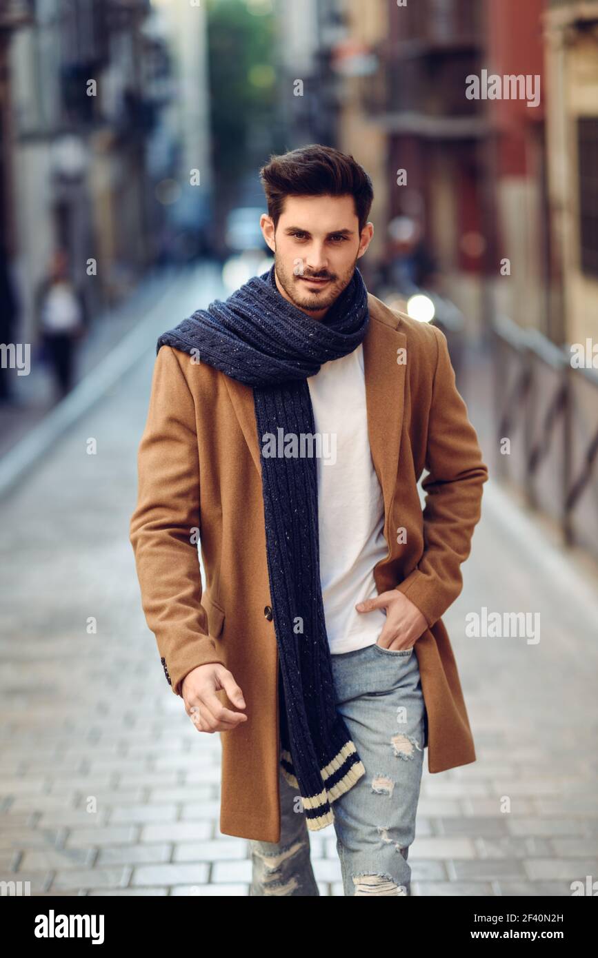 Hombre joven con ropa de invierno en la calle. Joven barbudo con peinado  moderno con abrigo, bufanda, jeans azules y camiseta. Hombre joven con ropa  de invierno en la calle Fotografía de