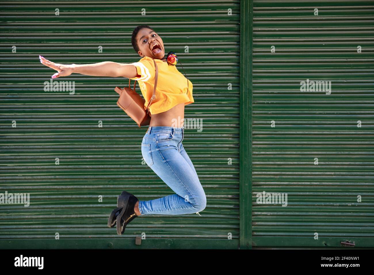 Joven mujer africana saltando sobre fondo de persianas. Chica feliz con pelo muy corto usando ropa informal. Joven mujer negra saltando sobre fondo de persianas. Foto de stock