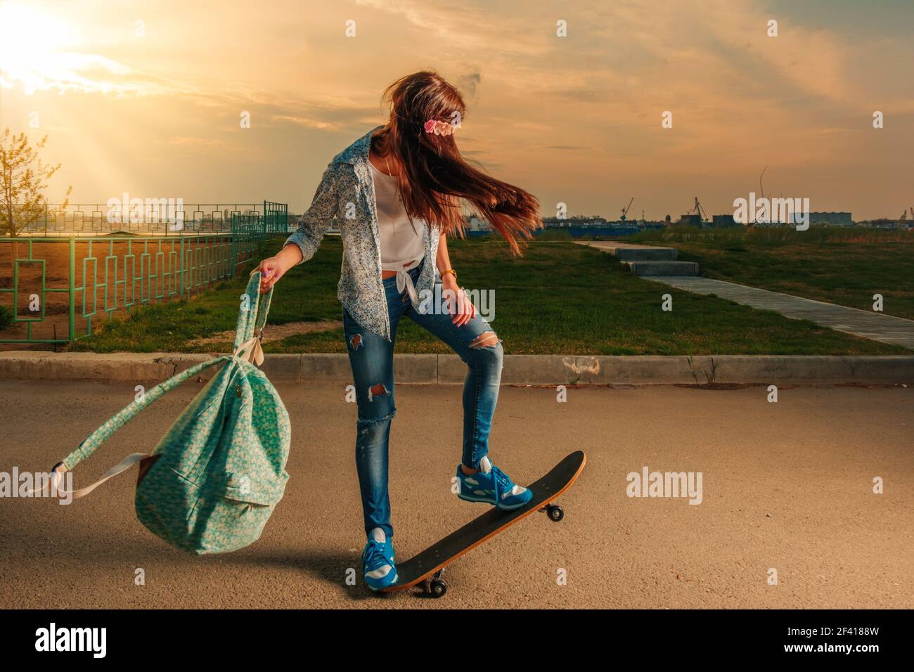 Hipster mujer en monopatín con mochila en sus manos retroiluminada en la  luz de la puesta de sol caliente haciendo truco en la carretera. Mujer  Hipster en monopatín con mochila en sus