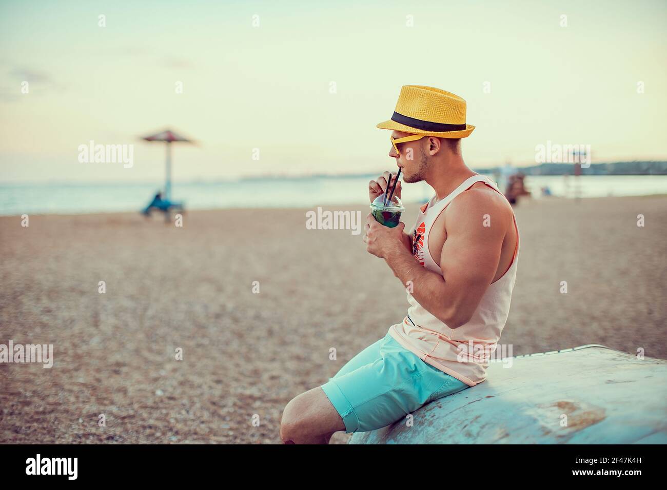 Joven hombre deportivo guapo con ropa brillante, sombrero de paja y gafas  de sol sentado en la orilla del mar, beber un refrescante cóctel y  disfrutar de sus vacaciones Fotografía de stock -