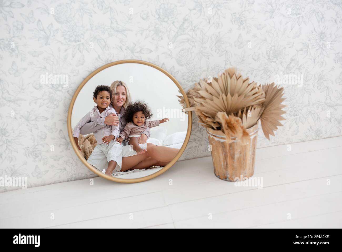 En el espejo de oro redondo, joven madre rubia caucásica sentada en suelo blanco de madera, con niños afroamericanos riendo. La mujer se abraza, besa hap Foto de stock
