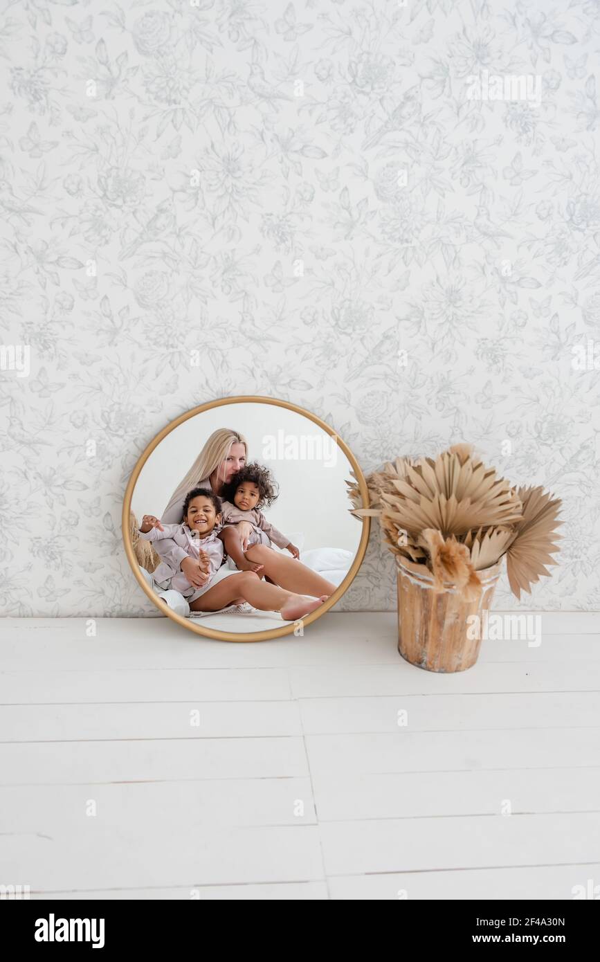 En el espejo de oro redondo, joven madre rubia caucásica sentada en suelo blanco de madera, con niños afroamericanos riendo. La mujer se abraza, besa hap Foto de stock