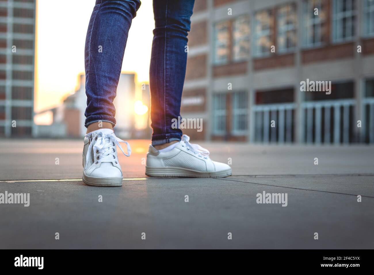 La mujer que lleva jeans y zapatillas de deporte está de pie en la ciudad  durante la puesta de sol. Enfoque selectivo. Moda y concepto elegante.  Zapatillas deportivas blancas al aire libre