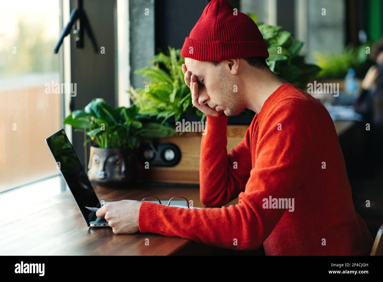 Hombre cansado tomando un descanso durante el trabajo en línea en el portátil, sosteniendo la cabeza en la mano, sintiendo falta de energía. Foto de stock