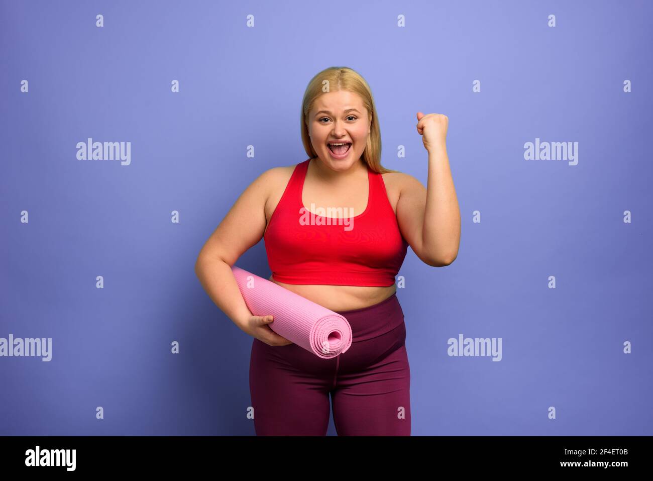 Chica gorda hace gimnasio en casa. Expresión satisfactoria y exitosa. Fondo púrpura. Foto de stock