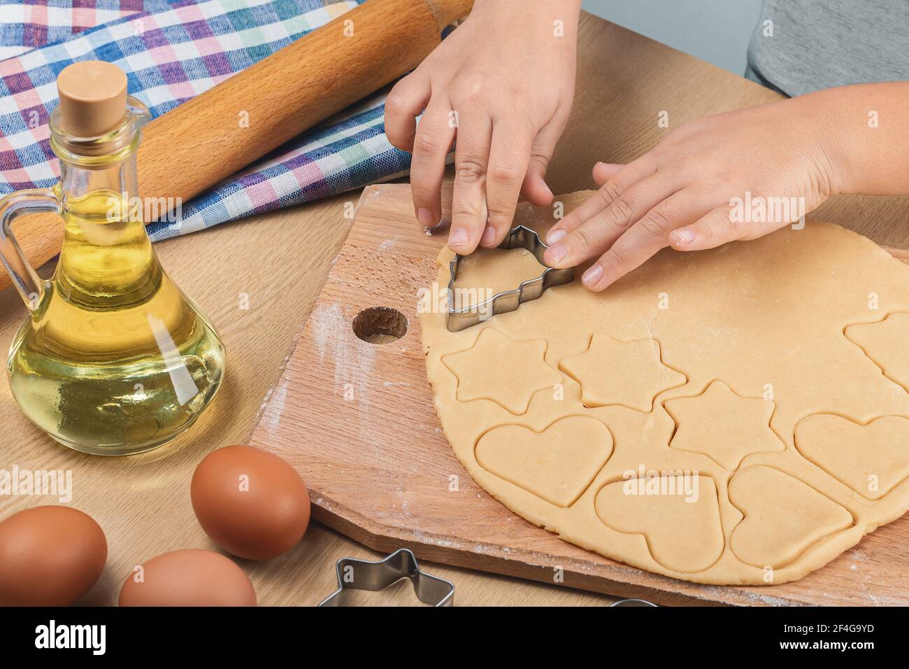 Las manos del niño sostienen un cortador de galletas de metal y cortan las  galletas caseras de varias formas de la masa de la corteza corta. En mesa  rodillo, huevos, girasol Fotografía