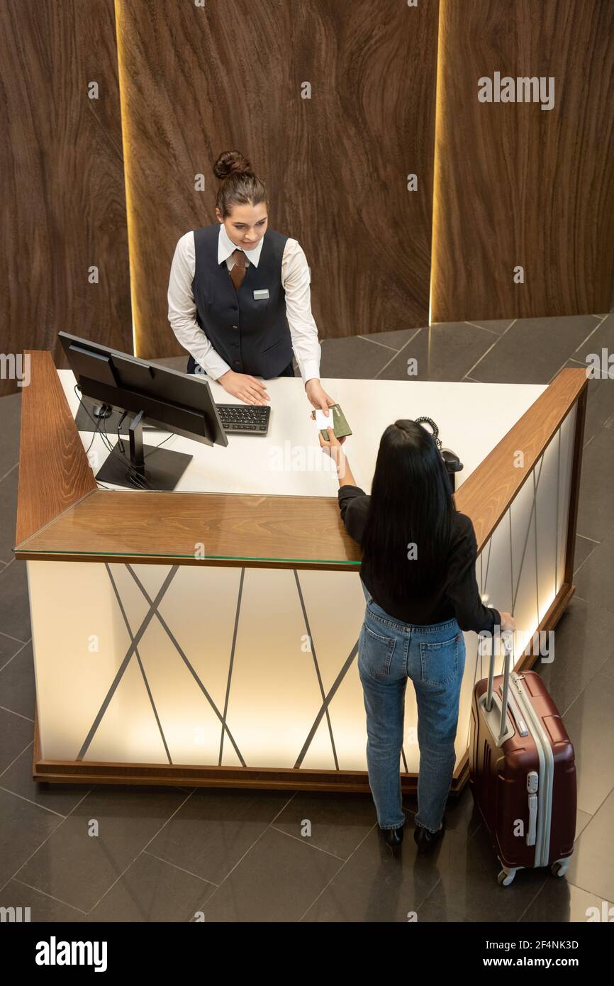 Joven recepcionista en uniforme parado por el lugar de trabajo y pasando el  pasaporte a la viajera femenina sobre el mostrador de recepción en el  vestíbulo de moderno hotel Fotografía de stock -