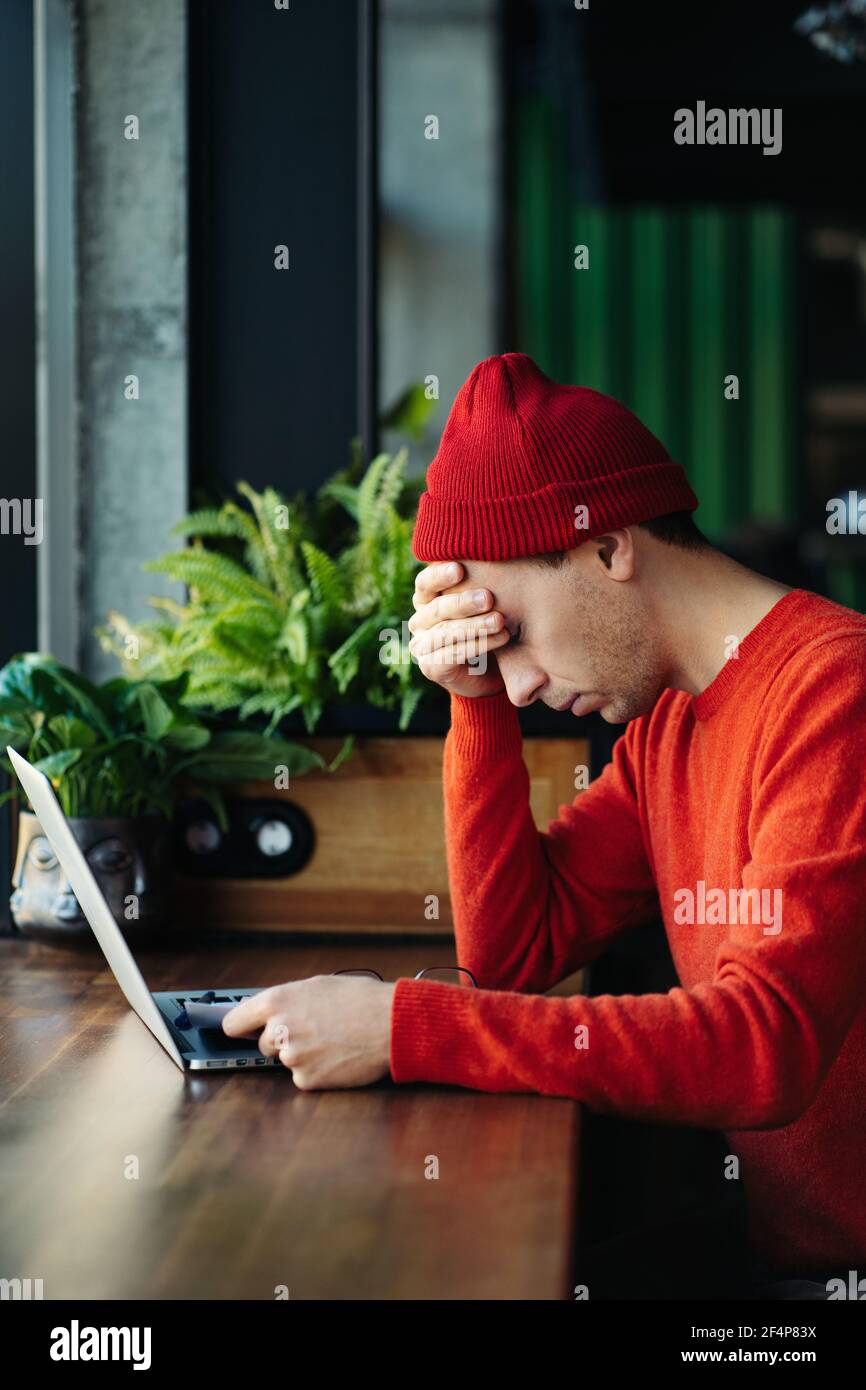 Hombre cansado tomando un descanso durante el trabajo en línea en el portátil, sosteniendo la cabeza en la mano, sintiendo falta de energía. Foto de stock
