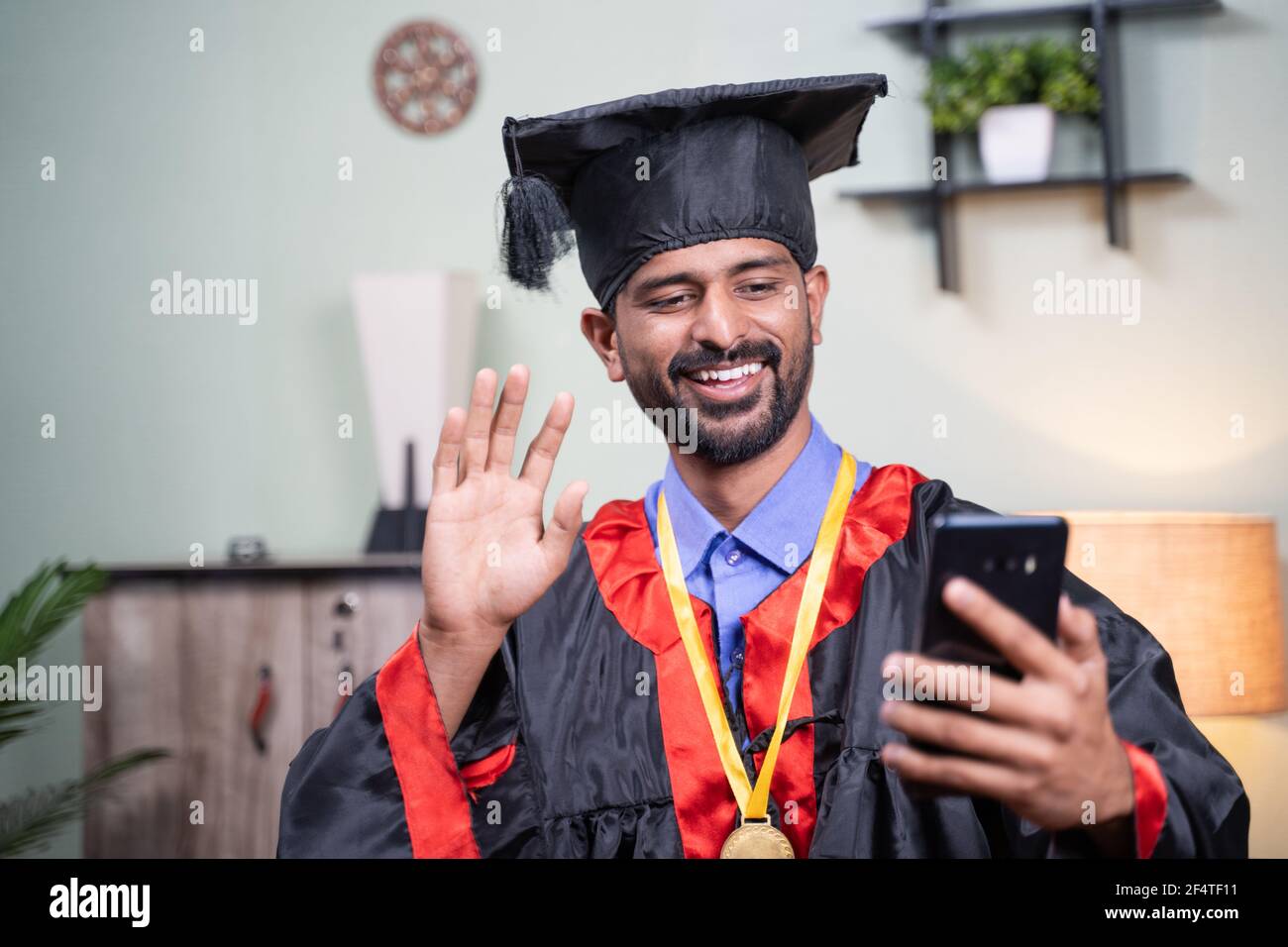 Estudiante que asiste a su celebración de graduación en vídeo en línea desde el teléfono móvil en el vestido de graduación - concepto de celebraciones virtuales, nuevo normal durante Foto de stock