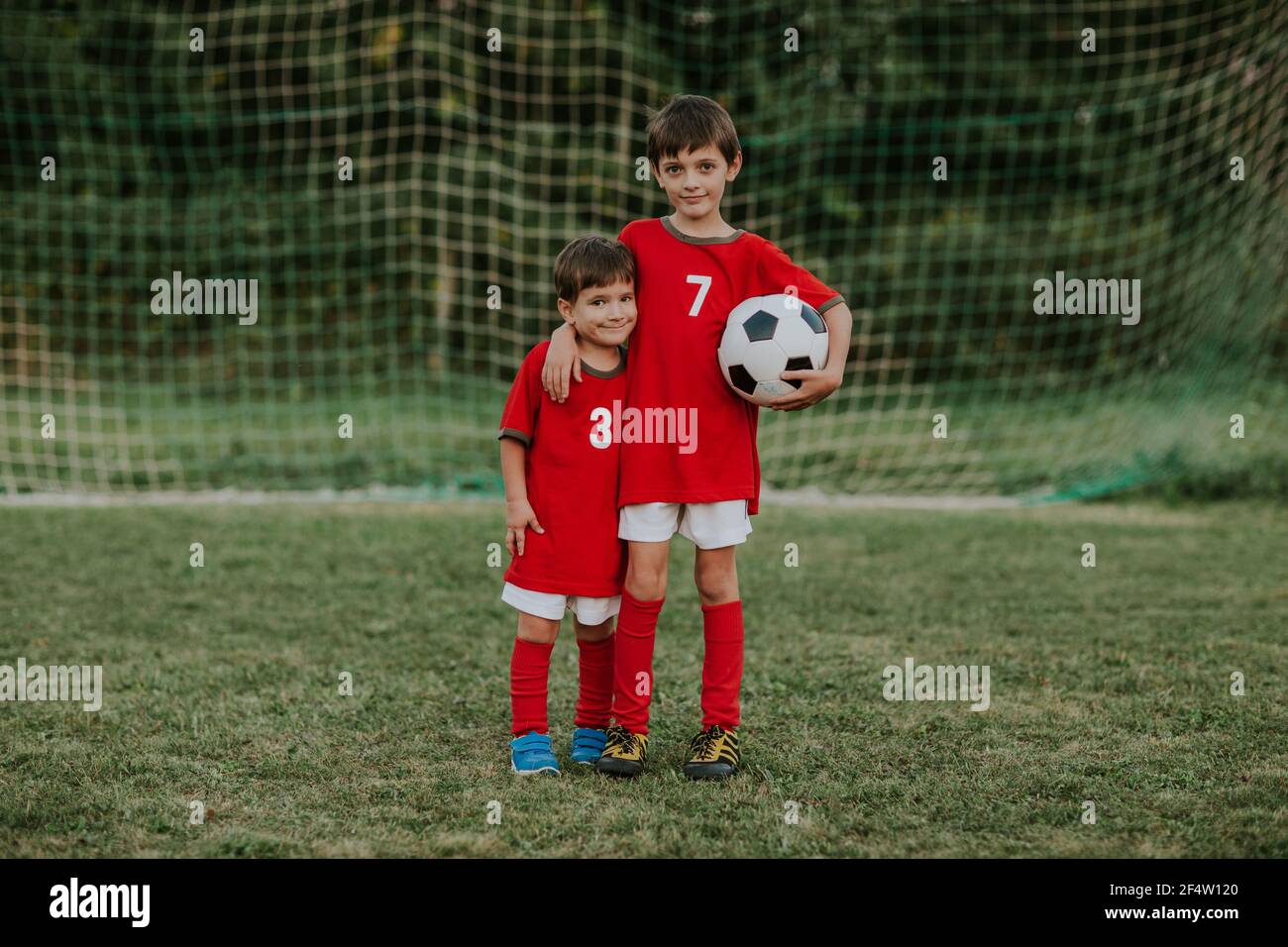 Jóvenes jugadores de fútbol de pie delante de la red de portería. Retrato completo de dos compañeros de equipo de fútbol con vestidos rojos sujetando el balón abrazándose unos a otros. Foto de stock